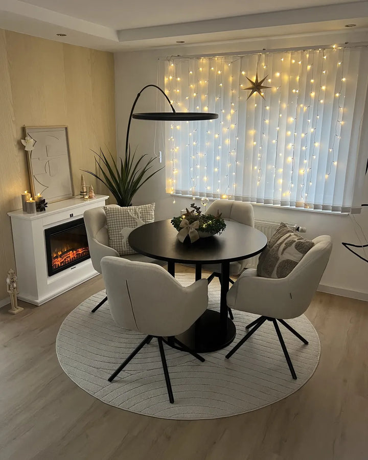 A modern dining area featuring a round black table surrounded by four cream upholstered chairs on a circular textured rug. The room is decorated with a white fireplace, a black floor lamp, and sheer curtains illuminated by warm string lights.