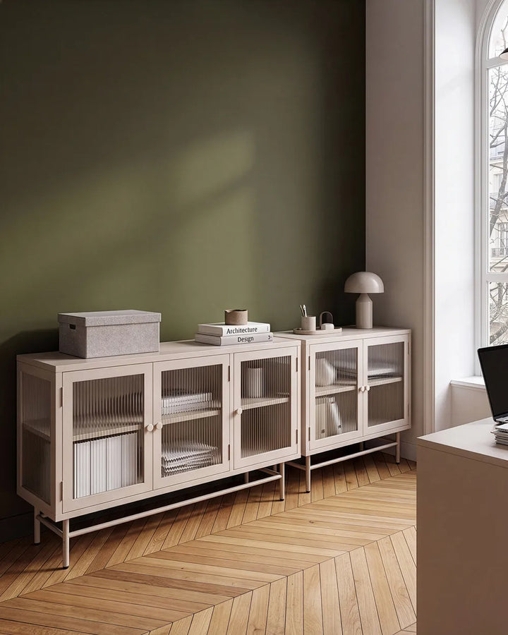 A light beige wooden sideboard cabinet with frosted glass doors in a modern room setting with a dark olive green wall and herringbone wood flooring.