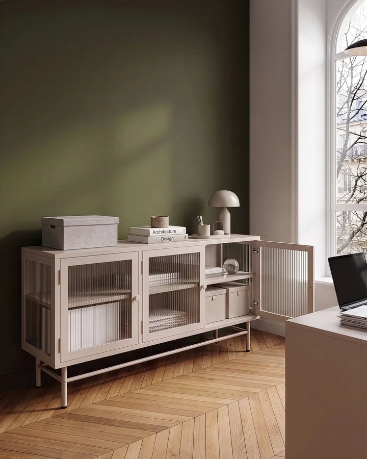 A light beige wooden sideboard cabinet with wire mesh doors and open shelving, styled in a minimalist room with a dark green wall, wood herringbone floor, and a window.