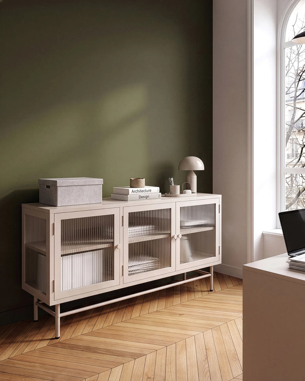 A modern sideboard cabinet with three glass-paneled doors and open lower shelves, styled in a minimalist room with a dark green wall, wood herringbone floor, and a window on the right.