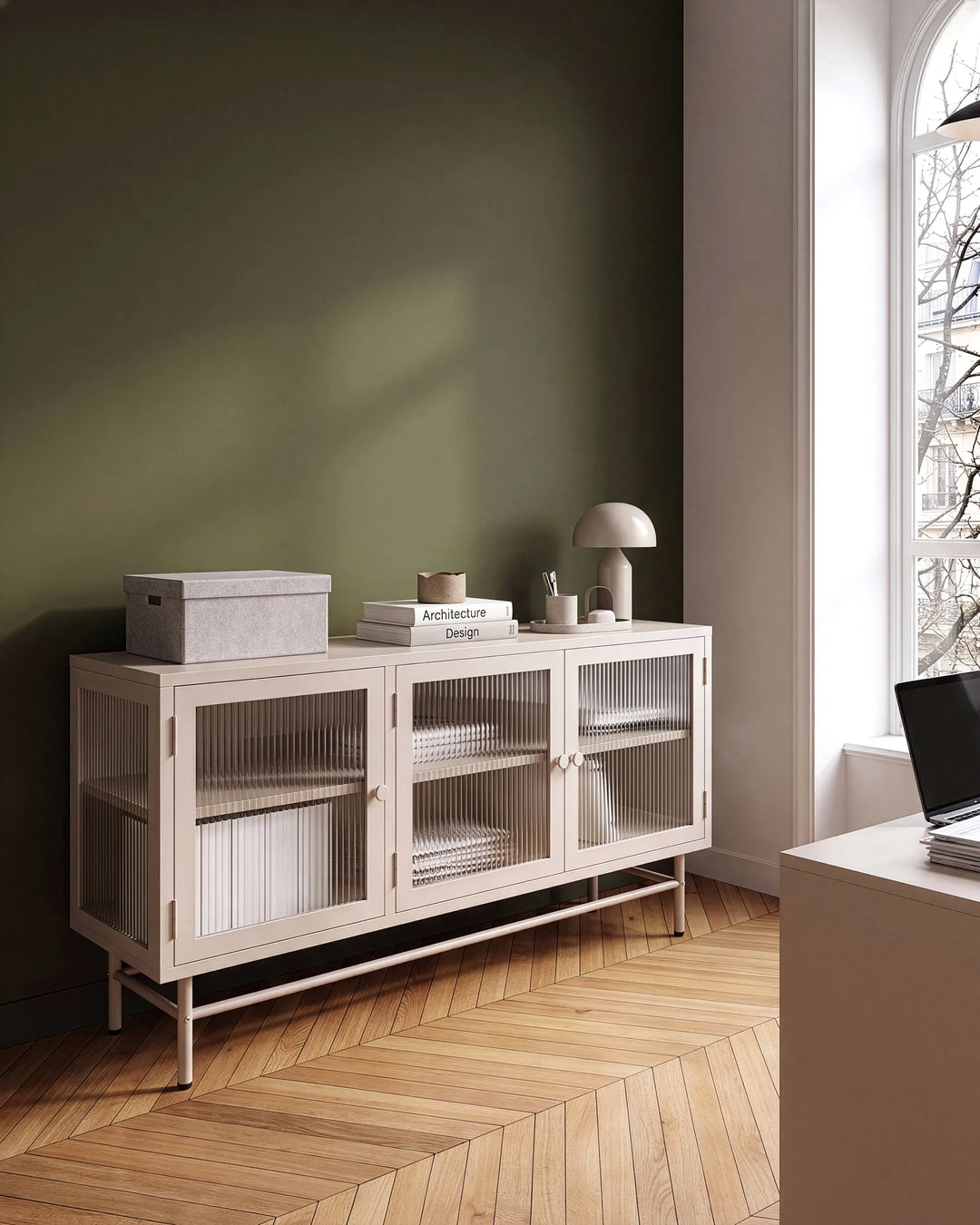 A modern sideboard cabinet with three glass-paneled doors and open lower shelves, styled in a minimalist room with a dark green wall, wood herringbone floor, and a window on the right.