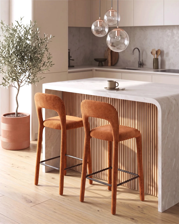 Two modern bar stools with orange-brown upholstered seats and backs, set beside a white kitchen island with a slatted wood front in a bright, contemporary kitchen.