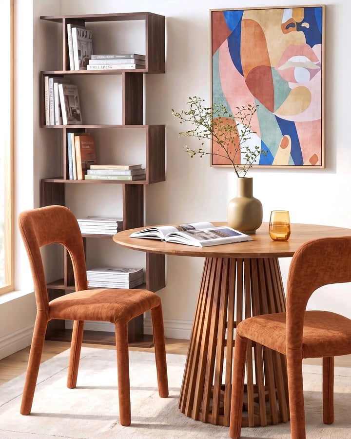 A styled dining area featuring a round wooden slatted table, two rust-orange upholstered chairs, a tall dark wood bookshelf, and a colorful abstract wall art print.