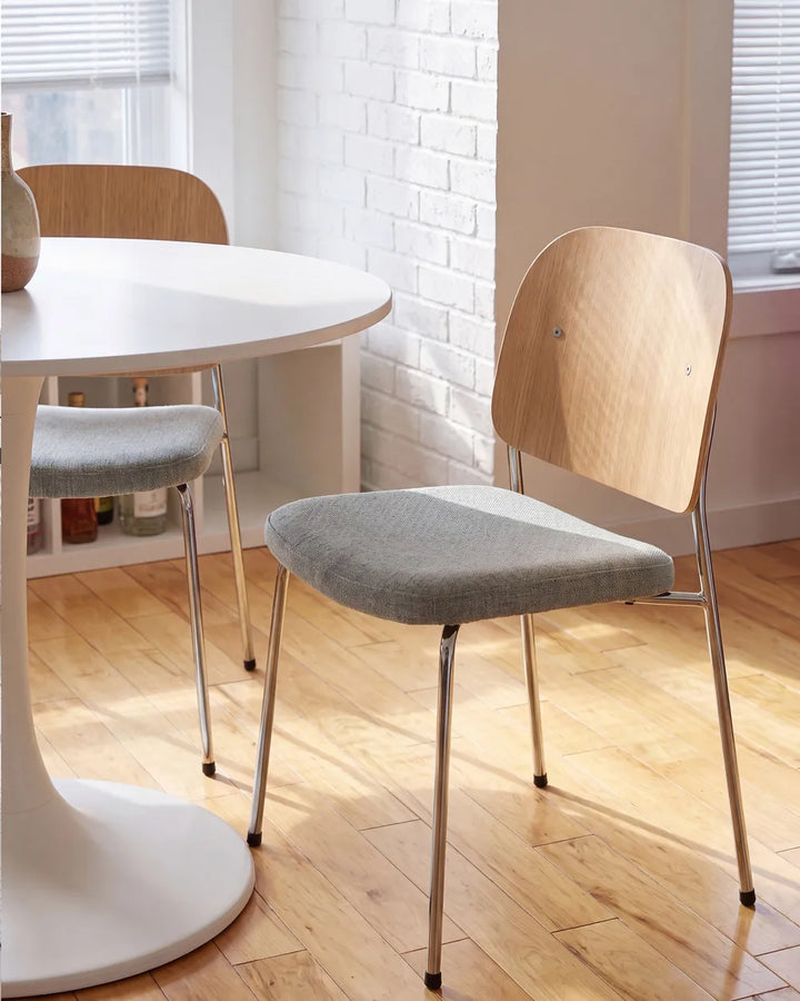 A modern dining chair with a light wood backrest and a gray upholstered seat, shown in a bright room with a white table, wood floor, and white brick wall.