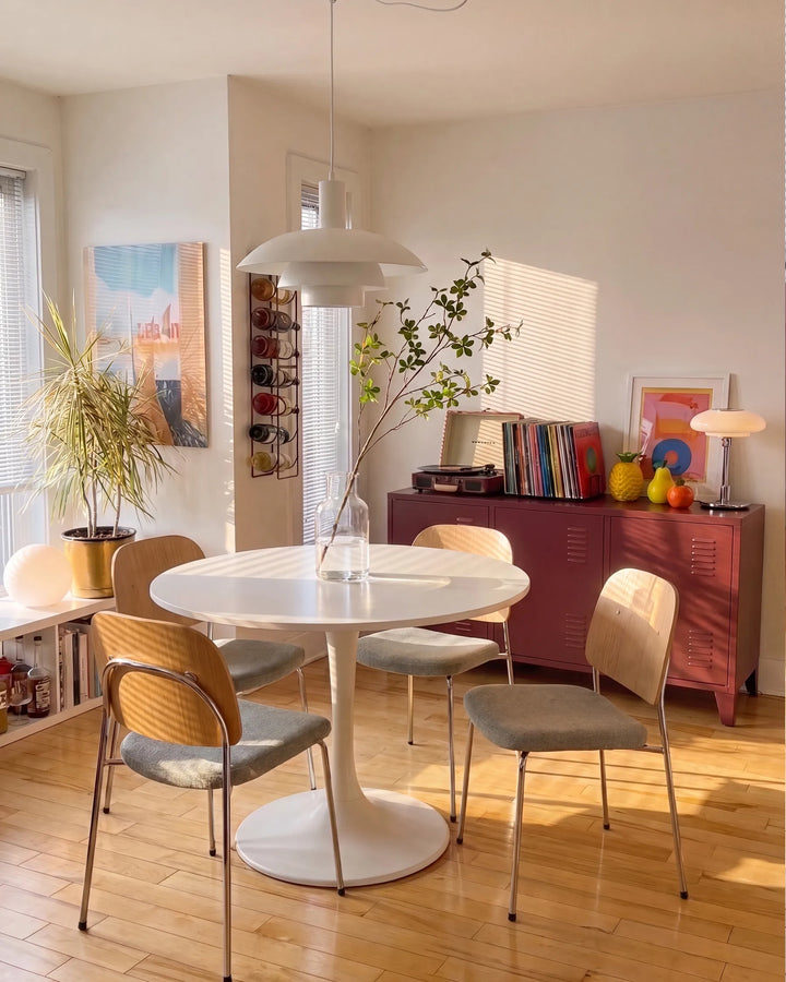 A modern dining room scene featuring a round white pedestal table surrounded by four mid-century style chairs with woven backs and gray upholstered seats. The room has light wood floors, a white pendant light, and a reddish-brown cabinet in the background.