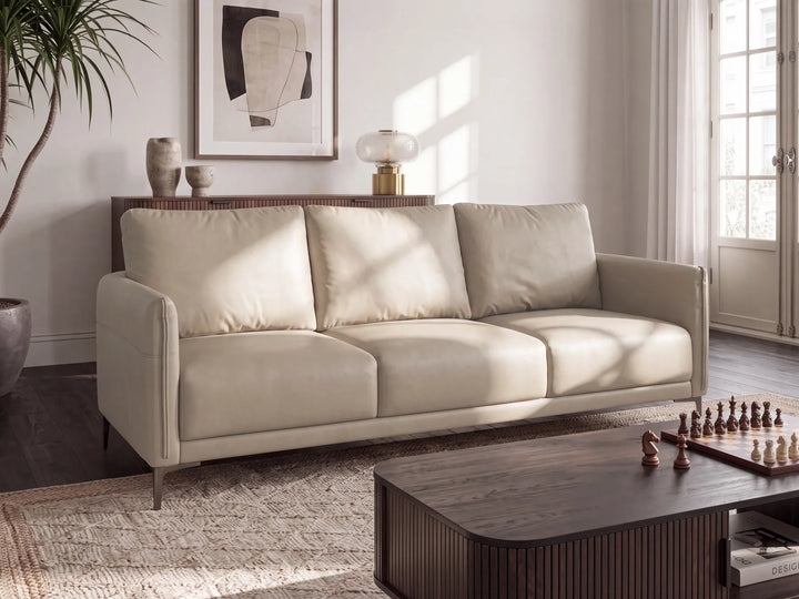 A modern three-seat sofa upholstered in light beige leather, styled in a bright, contemporary living room with a dark wood coffee table, patterned rug, and warm natural light.