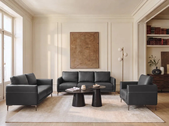 A modern living room set featuring a dark gray leather three-seater sofa, a matching two-seater armchair, and a single armchair, arranged around a round black coffee table on a light beige rug. The room has cream-colored walls with decorative molding, a large abstract painting, and built-in bookshelves.
