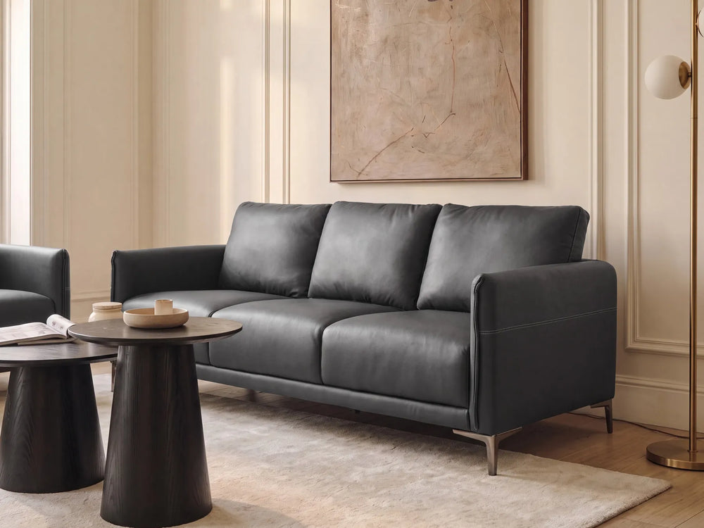 A modern three-seat sofa upholstered in dark charcoal gray leather, styled in a warm, elegant living room with cream walls, a beige rug, and dark wood tables.