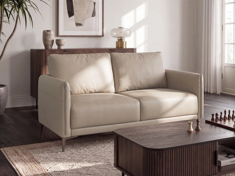 A modern two-seat sofa in light beige leather, styled in a contemporary living room with a dark wood coffee table, patterned rug, and wall art.