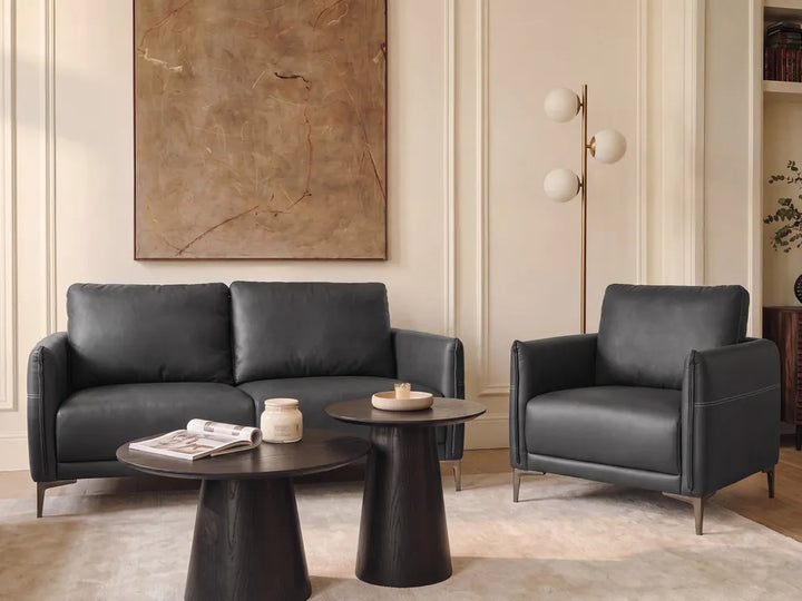 A modern living room setup featuring a black leather sofa and matching armchair with dark round coffee tables, set against cream paneled walls with a large abstract painting.
