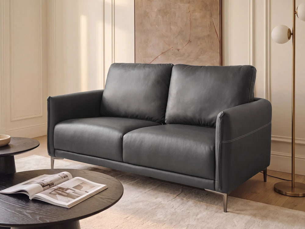 A modern two-seater sofa in dark charcoal gray leather, styled in a warm, elegant living room with cream walls, a marble wall art piece, a floor lamp, and round coffee tables.
