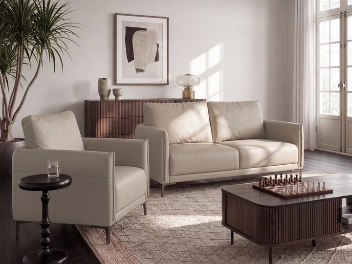 A modern living room setup featuring a light beige leather loveseat and matching armchair, a dark wood coffee table with a chess set, a patterned area rug, and a tall potted plant against a white wall.