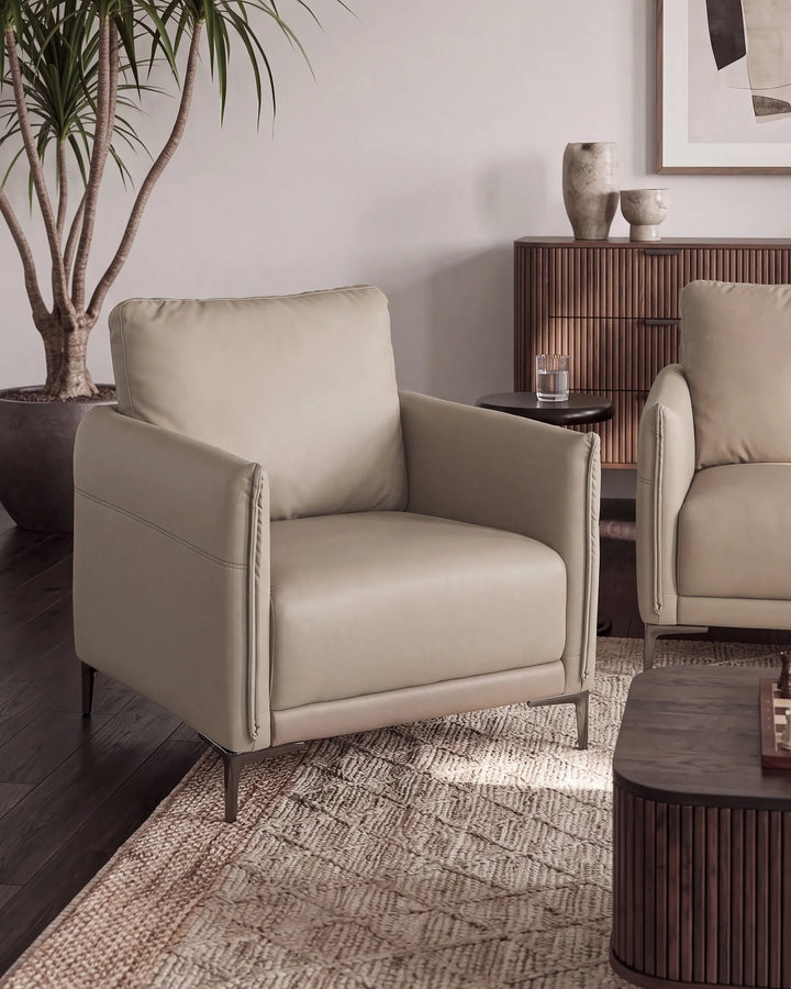 A modern beige leather armchair with dark metal legs, styled in a contemporary living room setting next to a matching armchair, a wooden sideboard, and a patterned rug.