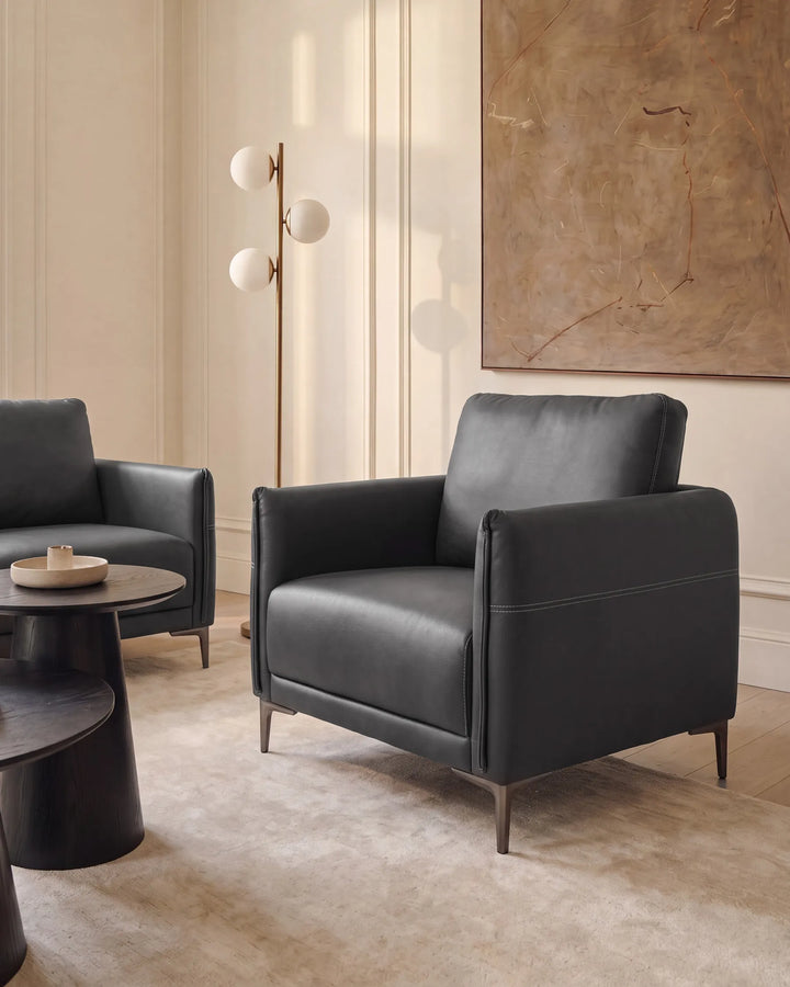 A modern black leather armchair with dark metal legs in a styled living room setting, featuring a matching armchair, round black coffee tables, a three-sphere floor lamp, and a large abstract painting on a cream paneled wall.