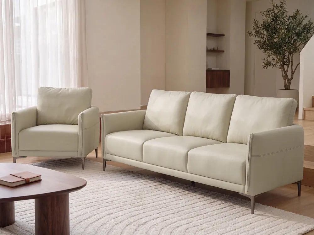 A modern cream-colored leather sofa and matching armchair set in a minimalist, warm-toned living room with a wooden coffee table and textured rug.