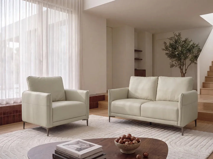A modern light beige leather armchair and matching two-seater sofa in a bright, minimalist living room with sheer curtains, a round coffee table, and a staircase in the background.