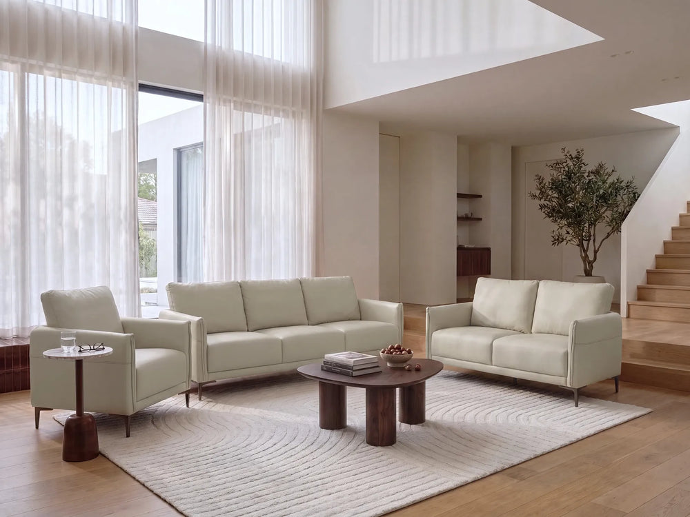 A modern living room featuring a light beige upholstered sofa, matching armchair, round dark wood coffee table, and a large textured cream rug on a light wood floor.