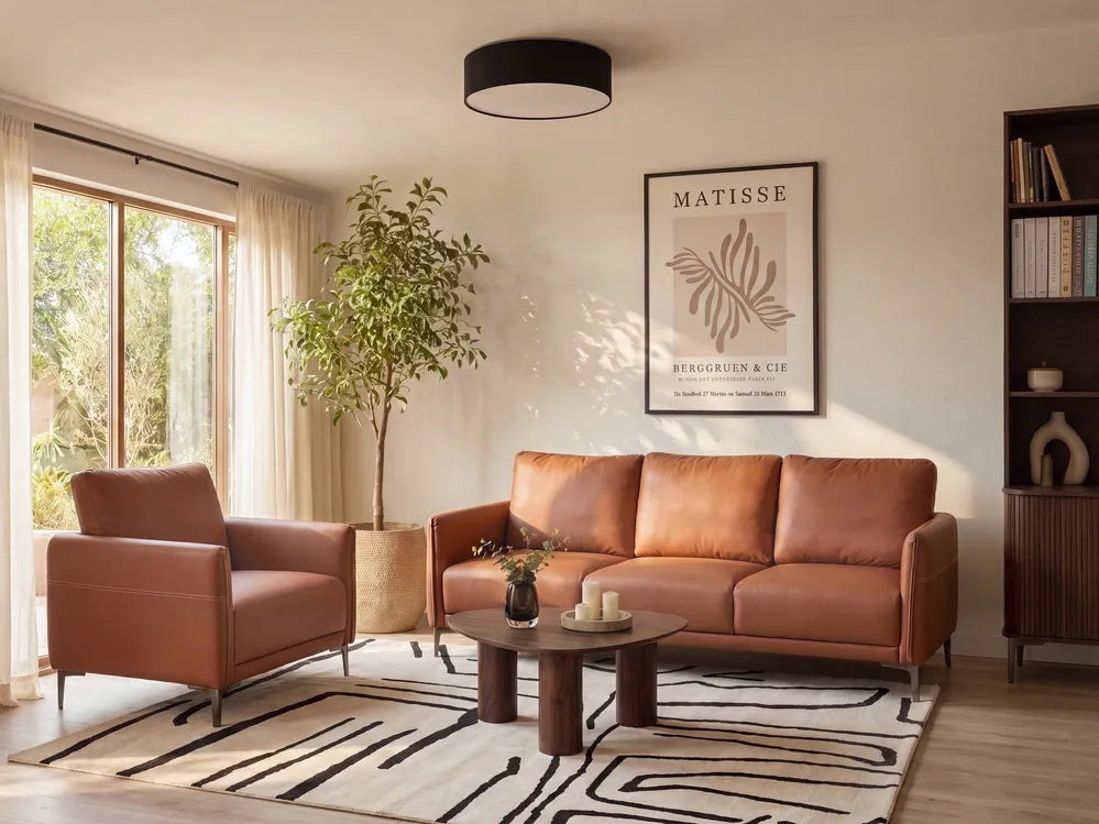 A warm brown leather sofa and matching armchair in a styled living room, with a round wooden coffee table, patterned rug, potted plant, and a framed Matisse print on the wall.
