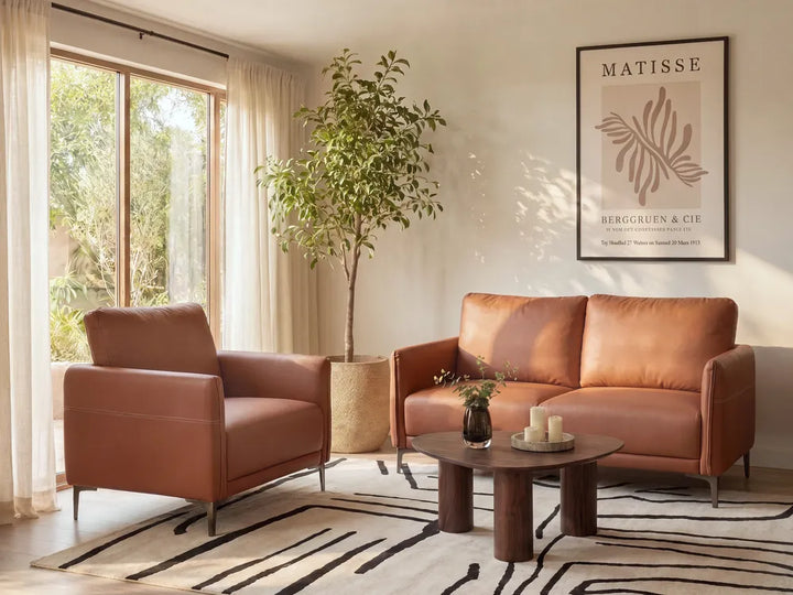 A warm brown leather armchair and matching loveseat arranged in a bright living room with a round wooden coffee table, patterned rug, potted tree, and framed Matisse wall art.