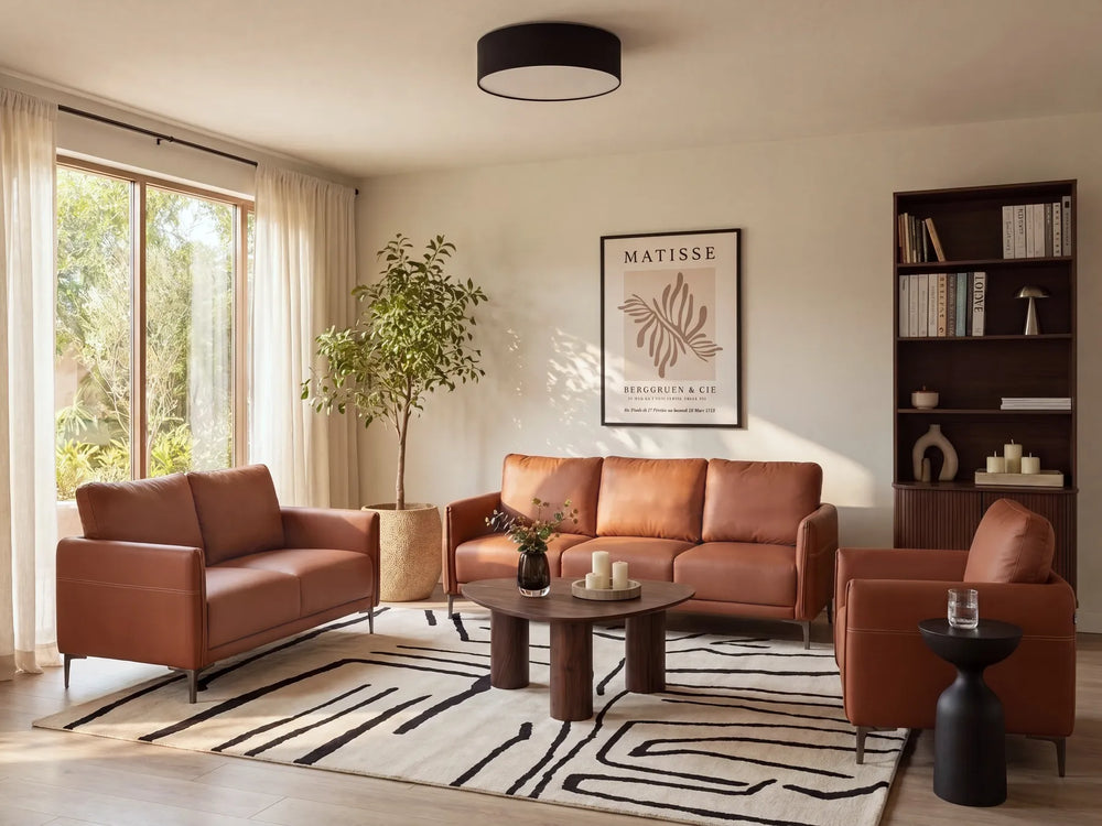 A styled living room featuring a rust-brown leather sofa, matching armchair, and a leather loveseat arranged around a round wooden coffee table on a black-and-white patterned rug. The room has beige walls, a large window with sheer curtains, a potted tree, a dark wood bookshelf, and a framed Matisse print.