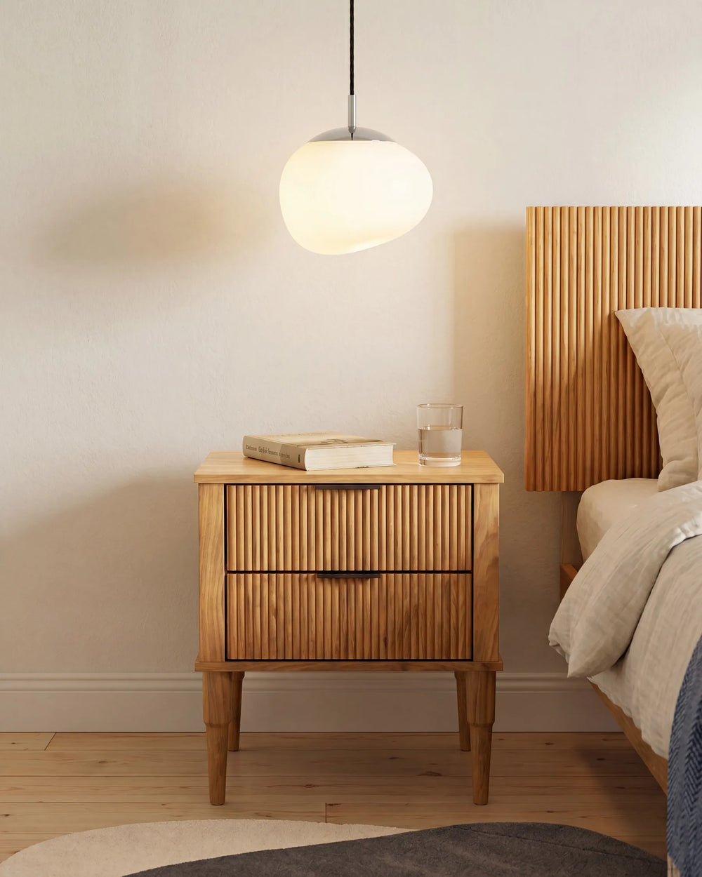 A mid-century modern wooden nightstand with slatted drawer fronts and tapered legs, styled in a bedroom next to a bed with a slatted wooden headboard. A glowing spherical pendant light hangs above, and a glass of water and a book sit on the nightstand.