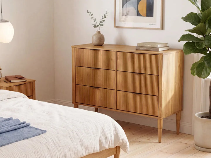 A mid-century modern wooden dresser with six vertically slatted drawers stands in a styled bedroom next to a bed, with a vase and books on top.