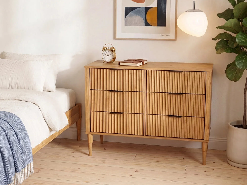 A mid-century modern wooden dresser with six drawers featuring vertical slat fronts and dark metal handles, styled in a bright bedroom next to a bed and a potted plant.