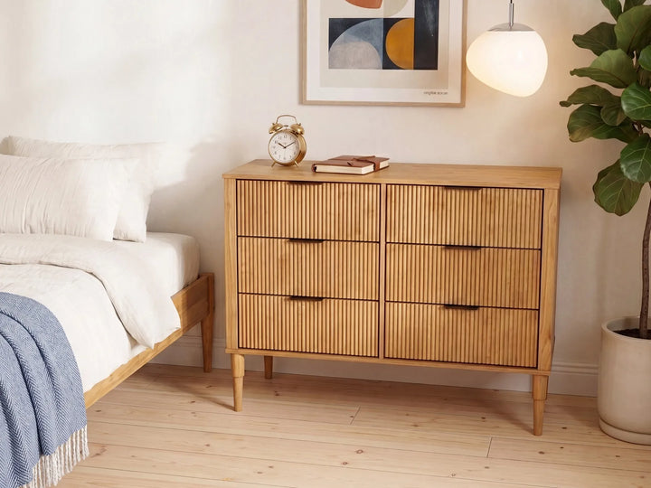 A mid-century modern wooden dresser with six drawers featuring vertical slat fronts and dark metal handles, styled in a bright bedroom next to a bed and a potted plant.