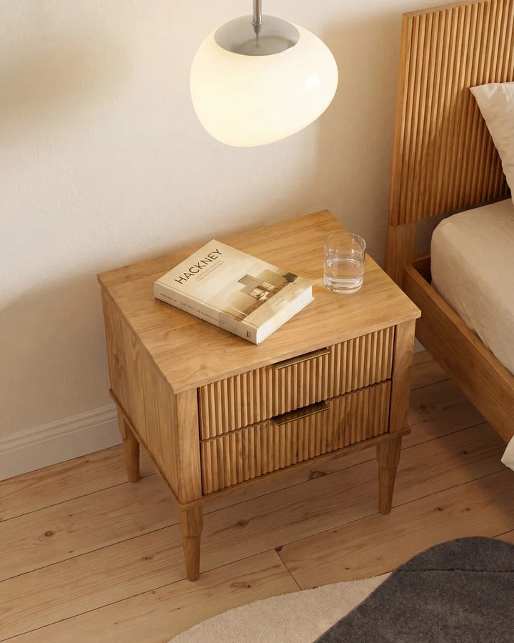 A light natural wood bedside table with two vertically slatted drawers, styled in a bedroom next to a matching wooden bed frame. A white spherical pendant lamp hangs above, and a book and a glass of water sit on the tabletop.