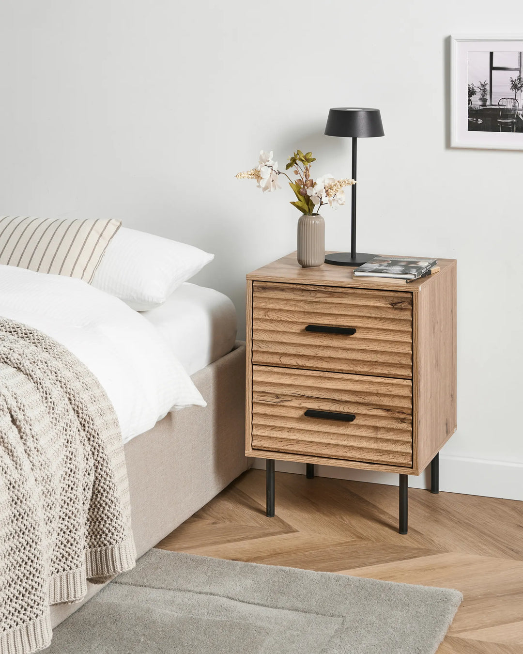 A two-drawer wooden nightstand with a slatted front and black metal legs, styled next to a bed in a modern bedroom setting.