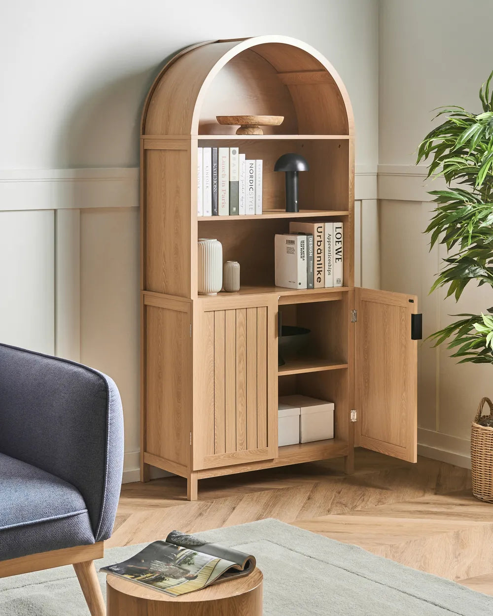 A tall light oak wood corner cabinet with an arched top, open upper shelves, two lower doors, and an open side door, styled in a living room corner next to a blue armchair and a potted plant.