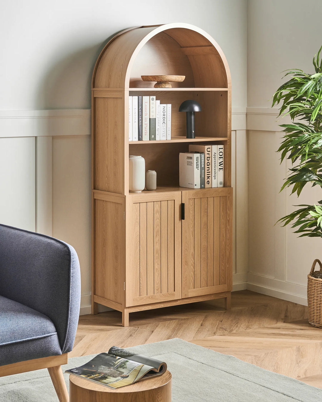 A tall light oak wood arched bookcase with three open shelves and two cabinet doors, styled in a cozy living room corner next to a blue armchair and a potted plant.