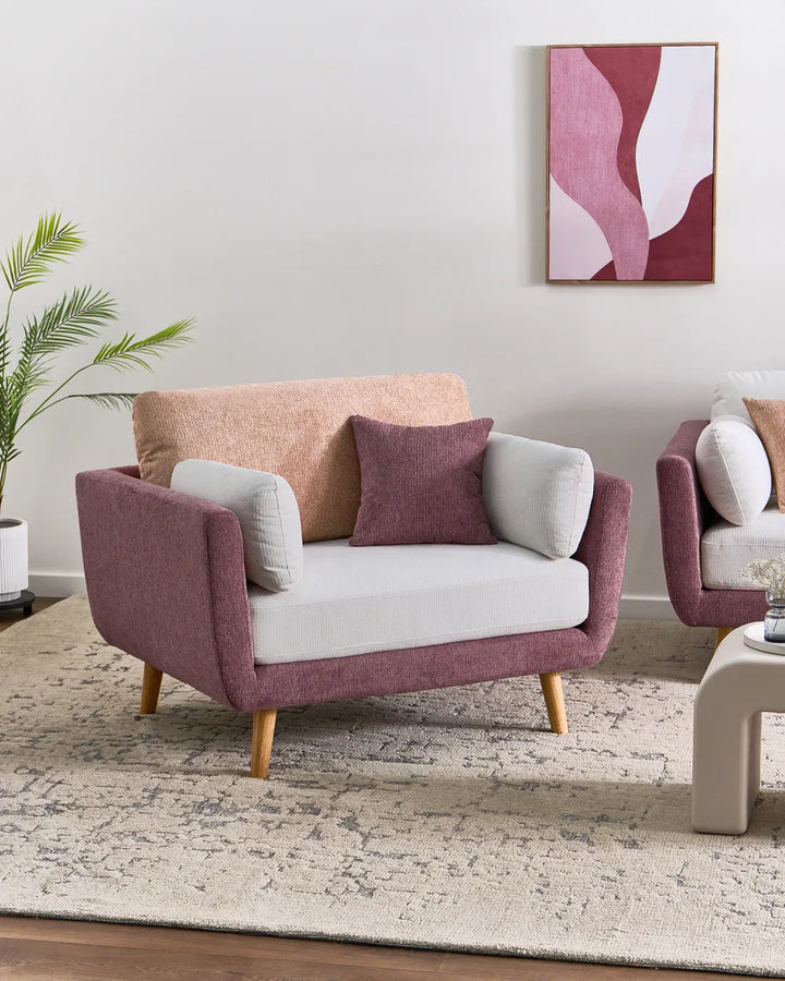 A modern loveseat sofa in dusty rose and white upholstery with light wood legs, styled in a living room setting with a beige rug, a potted plant, and abstract wall art.