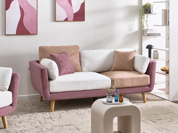 A modern three-seat sofa in a styled living room, featuring a dusty rose purple fabric frame, white and beige textured cushions, and light wood legs. The sofa is styled with matching accent pillows and is set against a white wall with abstract art.