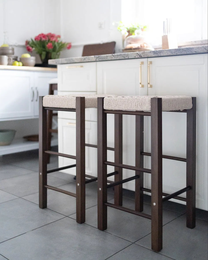 Two wooden bar stools with cream upholstered seats, positioned in front of a white kitchen island with a gray stone countertop.