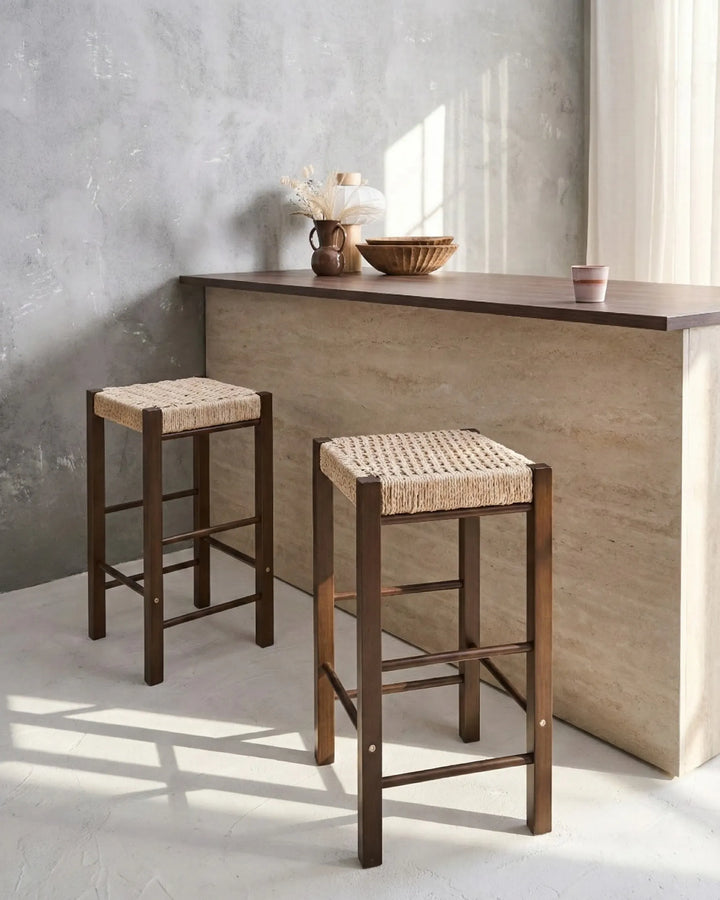 Two wooden bar stools with woven seats in front of a concrete wall and wooden counter.