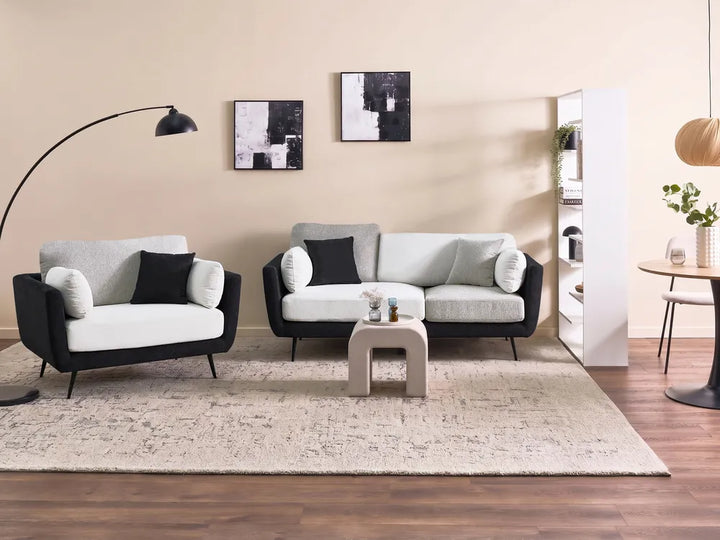 A modern living room setup featuring a black-framed two-seater sofa and matching armchair with light gray and white upholstery, styled on a large beige rug with a small side table in front.