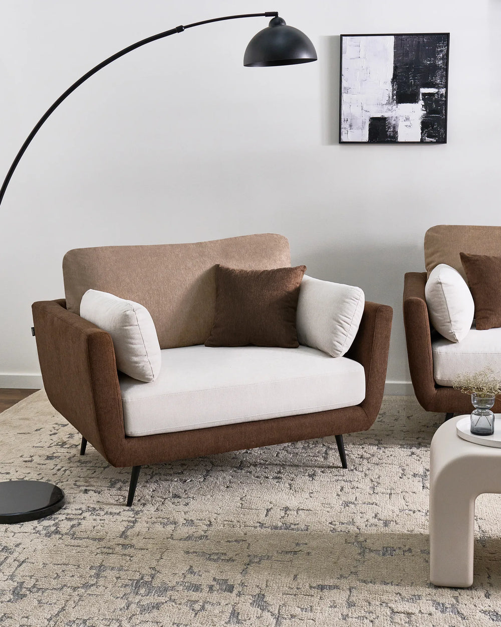 A modern two-seater armchair with a brown upholstered frame, cream seat cushion, and two accent pillows, styled in a minimalist living room setting with a matching armchair, floor lamp, and rug.