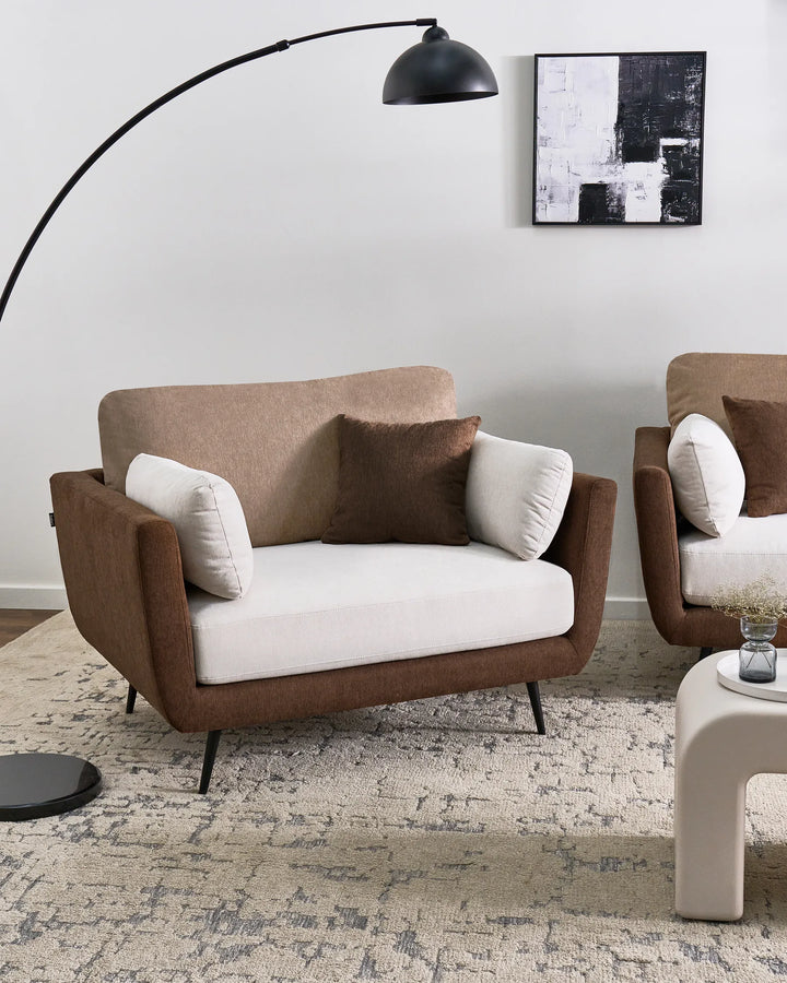 A modern two-seater armchair with a brown upholstered frame, cream seat cushion, and two accent pillows, styled in a minimalist living room setting with a matching armchair, floor lamp, and rug.