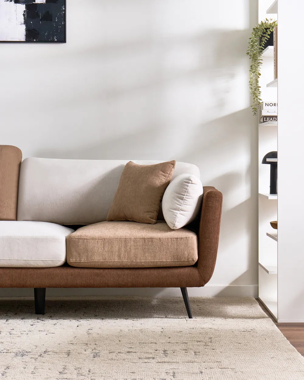 A modern three-seater sofa with a brown upholstered frame and mixed cream and taupe cushions, styled in a minimalist living room setting with a textured rug and a white shelving unit.