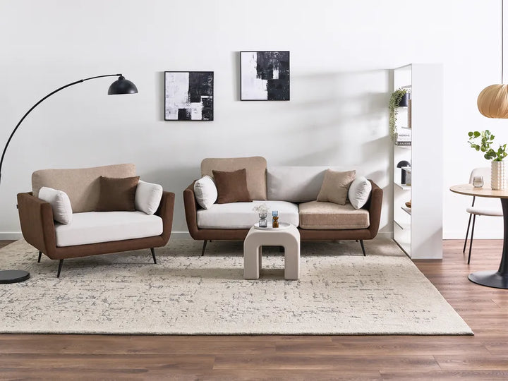 A modern living room furniture set featuring a three-seat sofa, a matching armchair, and a small concrete coffee table on a textured cream rug. The furniture has light beige upholstery, brown leather-look frames, and is styled in a bright, contemporary room with a white wall, wood floor, and a black floor lamp.