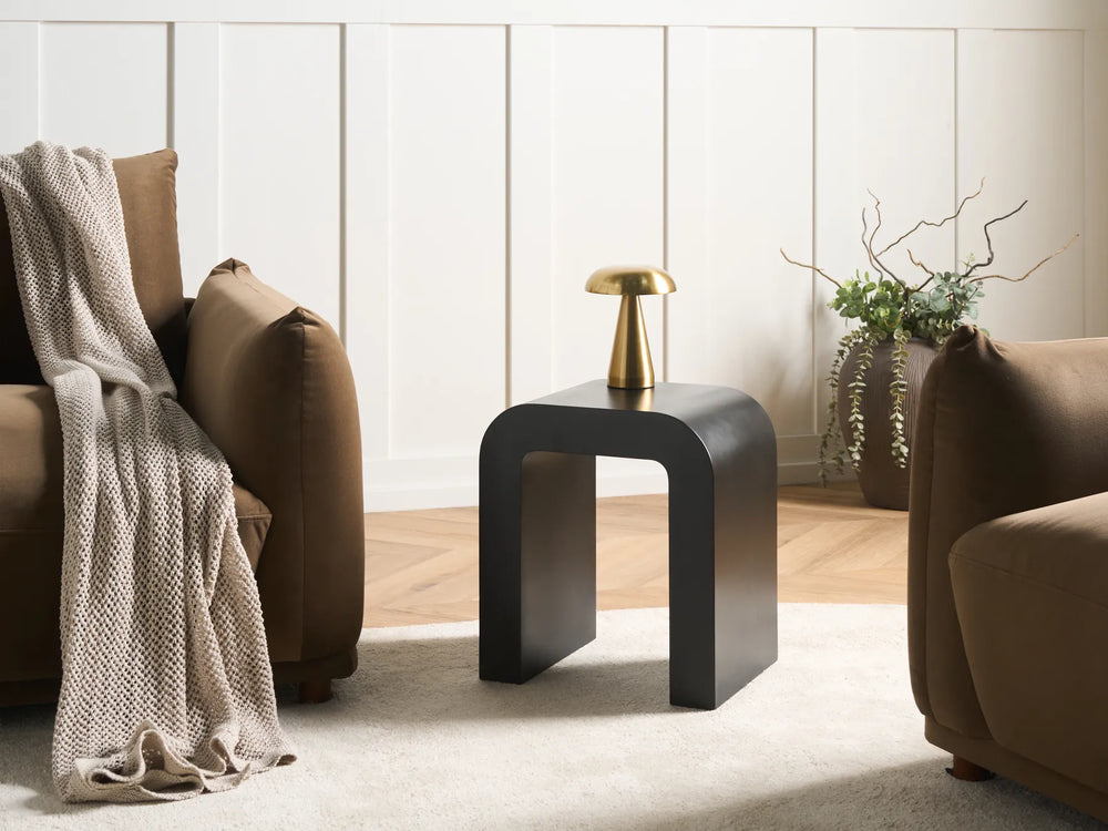 A modern black side table with a curved, open-frame design, styled in a living room between two brown armchairs with a beige throw blanket and a small gold table lamp on top.
