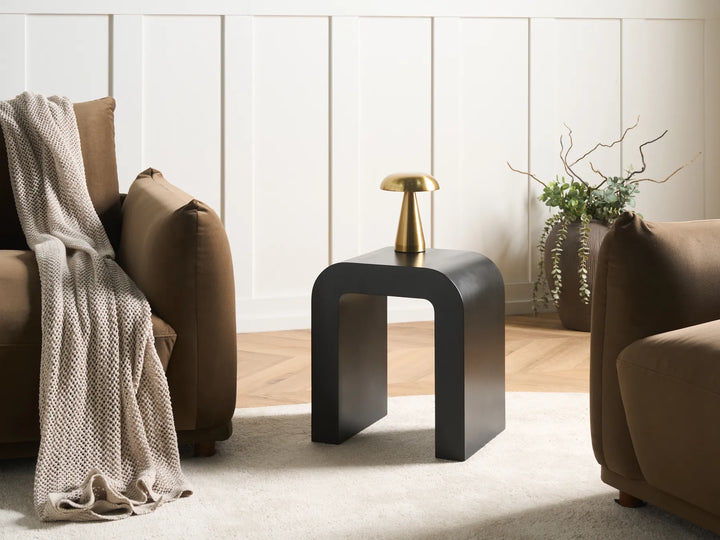 A modern black side table with a curved, open-frame design, styled in a living room between two brown armchairs with a beige throw blanket and a small gold table lamp on top.