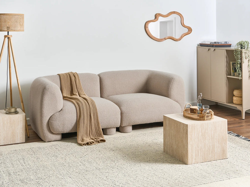 A modern curved two-seater sofa upholstered in a light beige bouclé fabric, styled in a minimalist living room with a textured beige throw blanket, a light wood cube side table, a woven floor lamp, and a matching cabinet against a white wall.