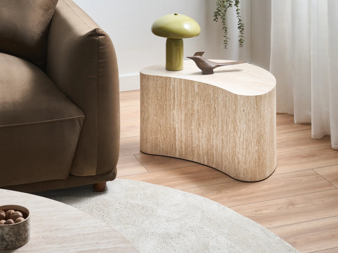 A modern organic-shaped wooden end table in a light natural finish, styled in a living room next to a brown sofa with a green mushroom lamp and a small bird figurine on top.