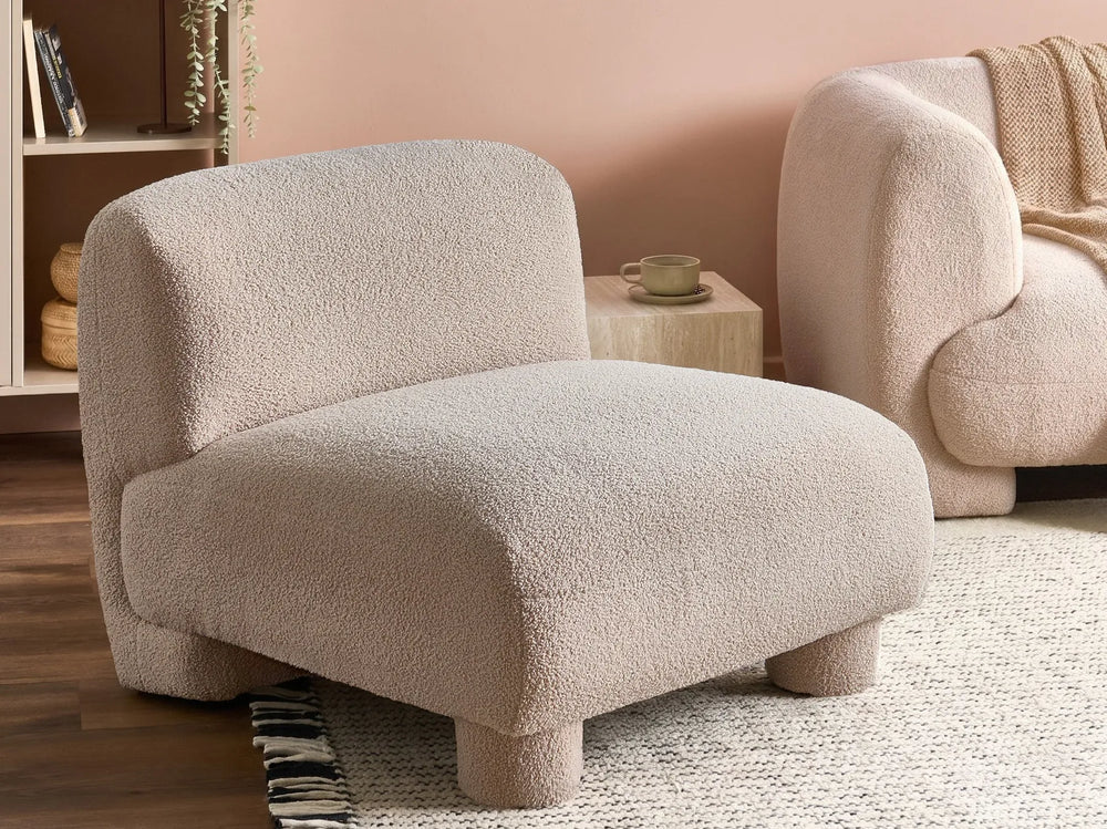 A modern low-profile accent chair upholstered in a textured boucle fabric in a warm light beige, set in a cozy living room next to a matching sofa.
