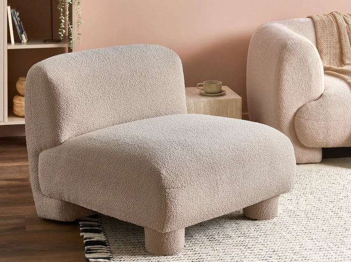 A modern low-profile accent chair upholstered in a textured boucle fabric in a warm light beige, set in a cozy living room next to a matching sofa.