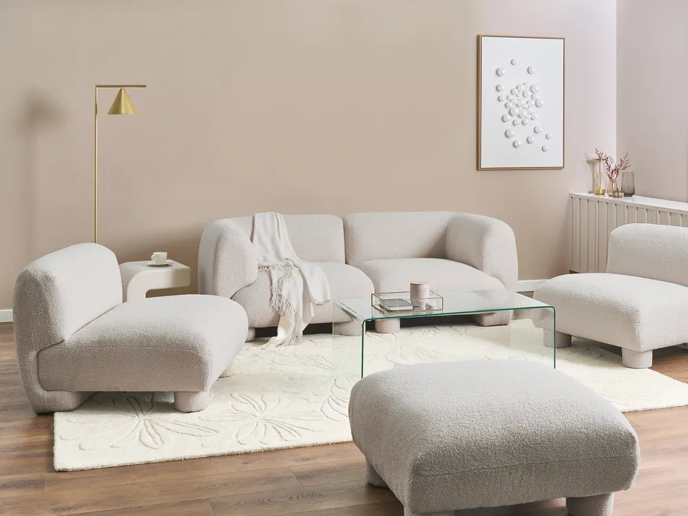 A modern living room set featuring a curved beige bouclé sofa, two matching armchairs, a large ottoman, a glass coffee table, and a textured cream rug on a wood floor against a beige wall.