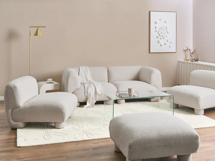 A modern living room set featuring a curved beige bouclé sofa, two matching armchairs, a large ottoman, a glass coffee table, and a textured cream rug on a wood floor against a beige wall.