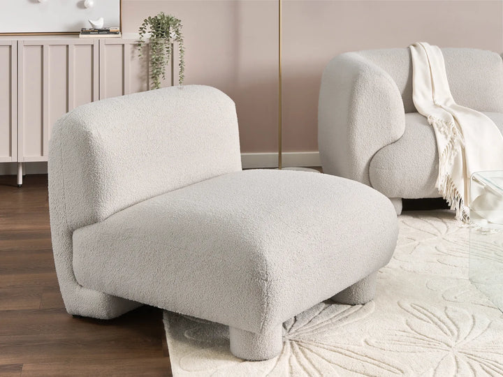 A modern low-profile accent chair upholstered in a bouclé fabric in a light warm gray or off-white color, styled in a minimalist living room next to another matching chair.